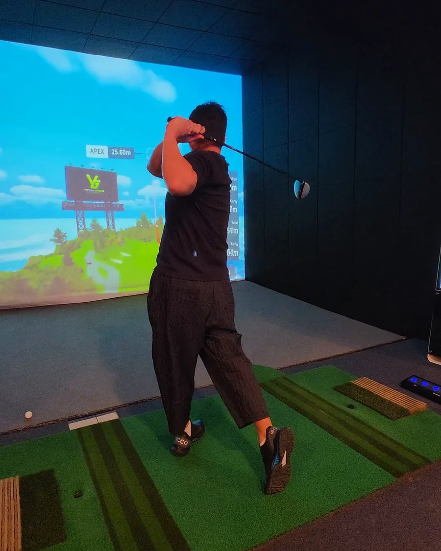 A golfer in black holding his follow-through in a Virtual Greens simulator bay, the VG board on the big screen